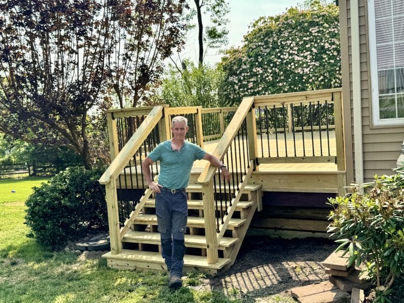 Man in teal shirt and gray pants poses on a newly built wooden outdoor staircase beside a house.