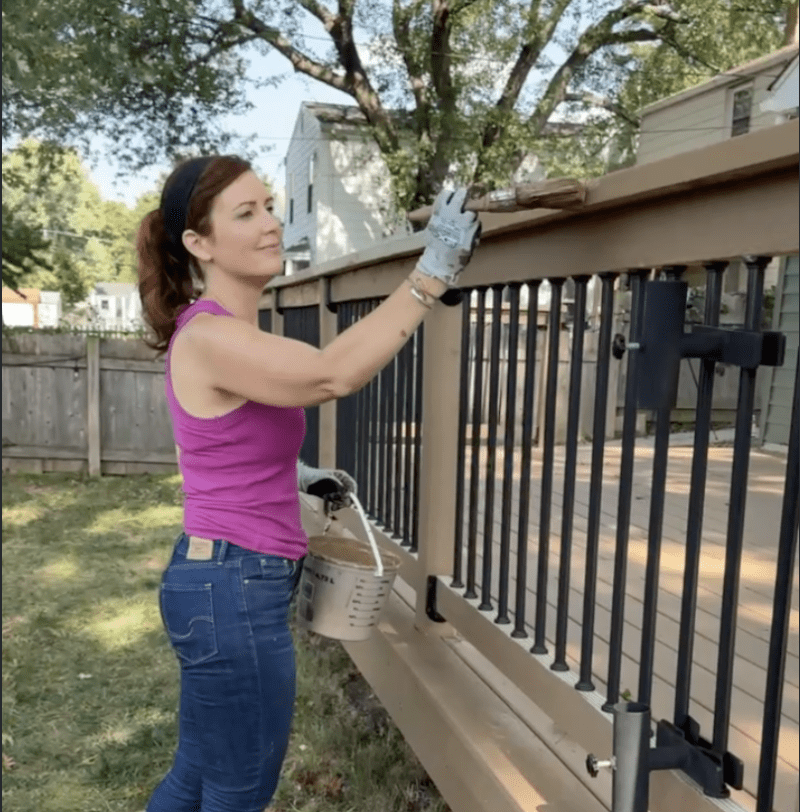 Woman in a pink tank top staining a wooden deck railing outdoors, holding a paintbrush and bucket nearby.