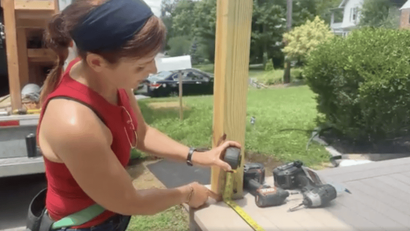 Woman in a red tank top measures a wooden post with a tape measure during outdoor construction of a deck or structure.